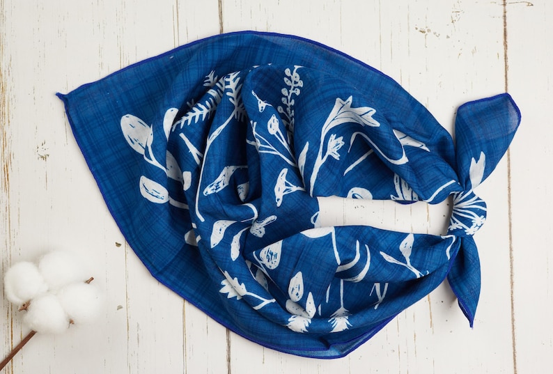 May include: A square, indigo blue scarf with a white floral pattern. The scarf is knotted and lies on a white wooden surface. The floral design includes various leaves and flower shapes. A cotton branch is in the lower left corner.