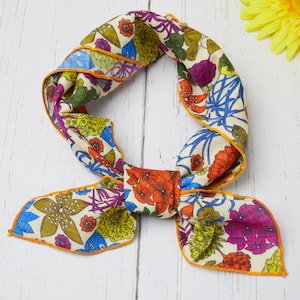 May include: A colorful floral scarf with a cream background, featuring orange, blue, purple, and green flowers. The scarf has an orange trim and is tied in a bow. The scarf is on a white wooden surface.