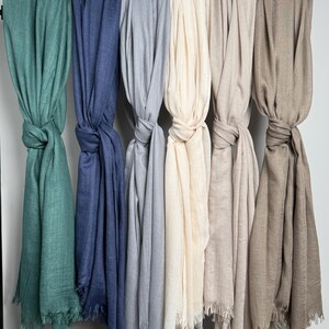 May include: A collection of six scarves in various colors, including teal, blue, gray, cream, beige, and brown. Each scarf is knotted and hangs from a rod, showcasing the fringed edges and soft texture of the fabric.