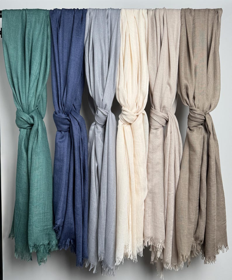 May include: A collection of six scarves in various colors, including teal, blue, gray, cream, beige, and brown. Each scarf is knotted and hangs from a rod, showcasing the fringed edges and soft texture of the fabric.