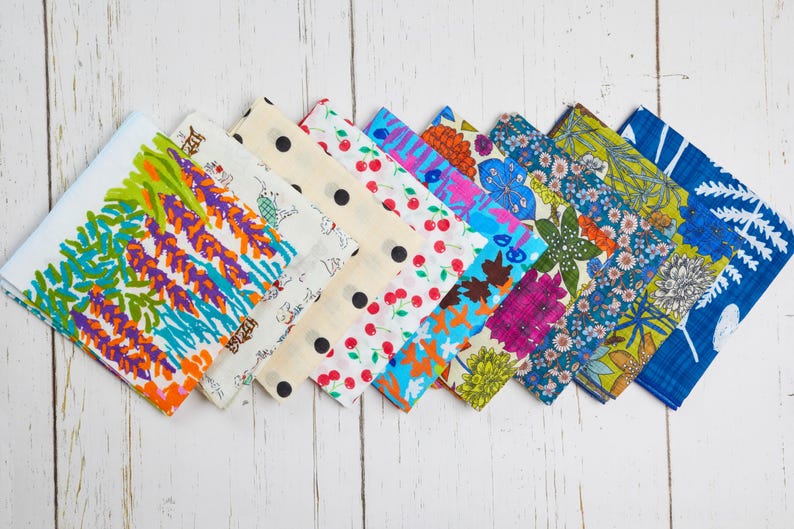 May include: A collection of colorful patterned fabric squares arranged diagonally. Designs include floral, polka dots, cherries, and abstract patterns in various colors like blue, green, orange, and pink. The fabric is displayed on a white wooden surface.