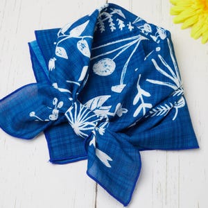 May include: A blue bandana with a white floral pattern is knotted on a white wooden surface. The bandana has a dark blue border and a lighter blue background. Yellow flowers are in the background.
