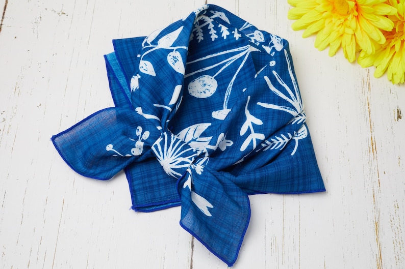 May include: A blue bandana with a white floral pattern is knotted on a white wooden surface. The bandana has a dark blue border and a lighter blue background. Yellow flowers are in the background.