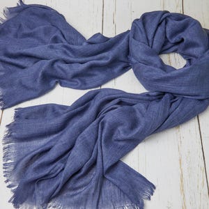 May include: A draped, denim blue scarf with fringed edges. The scarf is made of a soft, lightweight material and is spread out on a light-colored wooden surface. The scarf's texture is visible, and it appears to be new.