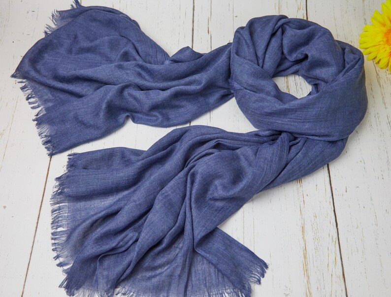 May include: A draped, denim blue scarf with fringed edges. The scarf is made of a soft, lightweight material and is spread out on a light-colored wooden surface. The scarf's texture is visible, and it appears to be new.