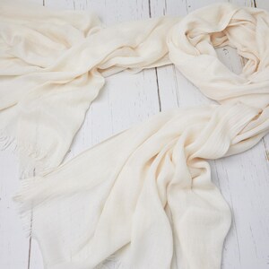 May include: A cream-colored scarf with fringed edges, draped on a light wood surface. The scarf is made of a soft, flowing fabric, with subtle texture and a light, neutral hue. The scarf is a versatile accessory.