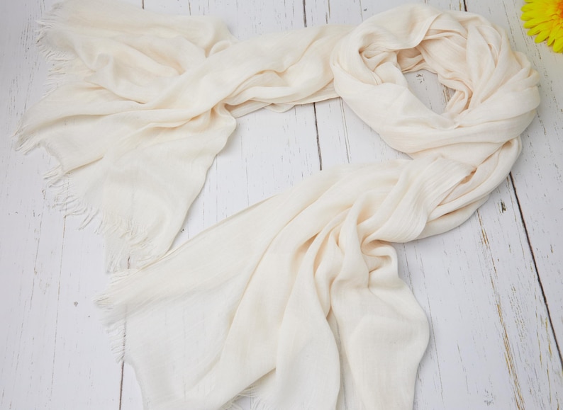 May include: A cream-colored scarf with fringed edges, draped on a light wood surface. The scarf is made of a soft, flowing fabric, with subtle texture and a light, neutral hue. The scarf is a versatile accessory.