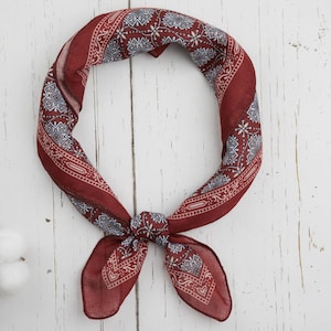 May include: A burgundy bandana with a paisley pattern in white and blue, tied in a knot. The fabric is folded and arranged in a circular shape, with the knot at the bottom. The bandana is on a white wooden surface.