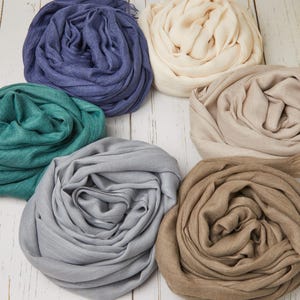 May include: A collection of six rolled scarves in various colors, including blue, teal, gray, beige, and brown. The scarves are arranged on a white wooden surface, with a yellow flower in the background. The scarves appear to be made of a soft, lightweight fabric.