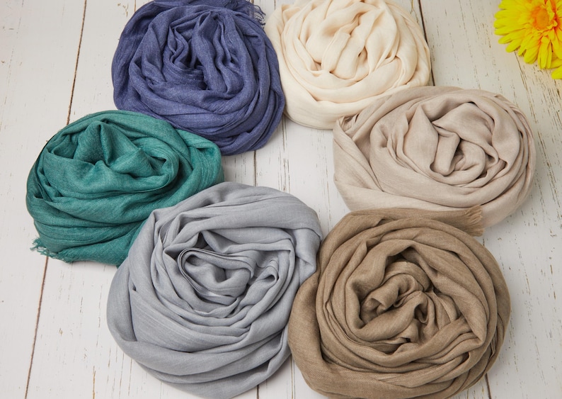 May include: A collection of six rolled scarves in various colors, including blue, teal, gray, beige, and brown. The scarves are arranged on a white wooden surface, with a yellow flower in the background. The scarves appear to be made of a soft, lightweight fabric.