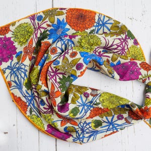 May include: A colorful floral scarf with a cream background and a vibrant array of flowers in orange, blue, pink, and green. The scarf has an orange border and is knotted at one end.