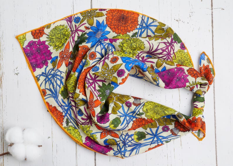 May include: A colorful floral scarf with a cream background and a vibrant array of flowers in orange, blue, pink, and green. The scarf has an orange border and is knotted at one end.