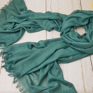 May include: A teal-colored scarf with fringed edges, draped on a light wood surface. The scarf is made of a soft, flowing fabric, with visible texture and a slightly wrinkled appearance. The scarf is a solid color and appears to be lightweight.