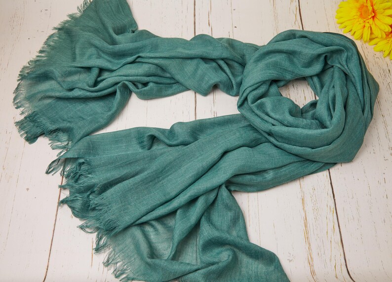 May include: A teal-colored scarf with fringed edges, draped on a light wood surface. The scarf is made of a soft, flowing fabric, with visible texture and a slightly wrinkled appearance. The scarf is a solid color and appears to be lightweight.