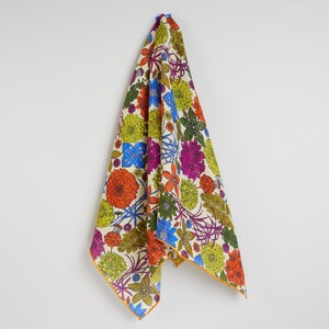 May include: A colorful floral patterned textile, featuring orange, blue, purple, and green flowers on a cream background. The edges are trimmed with a thin yellow border. The fabric is hanging against a white wall.