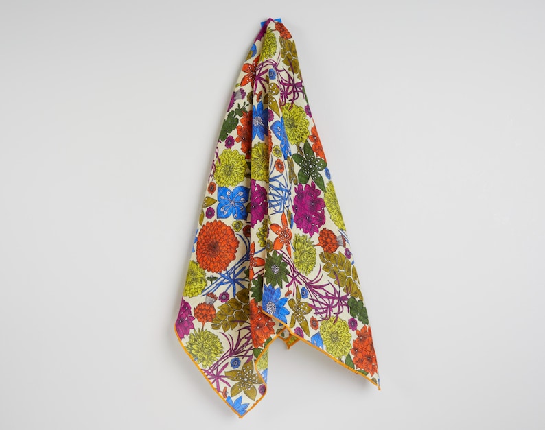 May include: A colorful floral patterned textile, featuring orange, blue, purple, and green flowers on a cream background. The edges are trimmed with a thin yellow border. The fabric is hanging against a white wall.