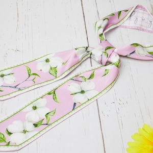 Puede incluir: Un pañuelo rosa con flores de cornejo blancas y hojas verdes. El pañuelo está anudado, con los extremos extendidos. El fondo es una superficie de madera blanca. Flores amarillas en la esquina inferior derecha.