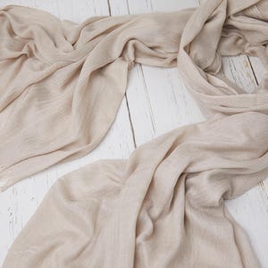 May include: A light beige scarf with fringed edges, draped on a white wooden surface. The scarf is made of a soft, flowing fabric, with visible texture and subtle variations in color. The scarf is a neutral accessory.