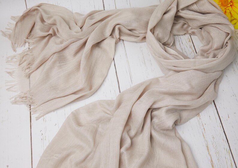 May include: A light beige scarf with fringed edges, draped on a white wooden surface. The scarf is made of a soft, flowing fabric, with visible texture and subtle variations in color. The scarf is a neutral accessory.