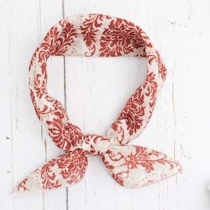 May include: A cream-colored bandana with a repeating red floral pattern, tied in a knot. The bandana is made of a lightweight fabric and is displayed on a white wooden surface. The overall aesthetic is vintage and feminine.