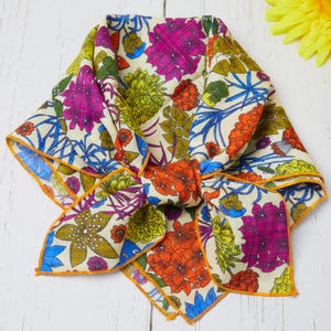 May include: A folded floral scarf with a cream background and a vibrant pattern of purple, orange, blue, and green flowers. The scarf has an orange border and is knotted in the center.