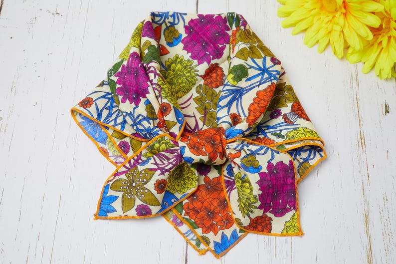 May include: A folded floral scarf with a cream background and a vibrant pattern of purple, orange, blue, and green flowers. The scarf has an orange border and is knotted in the center.