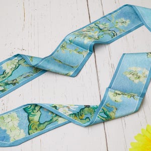 Puede incluir: Un pañuelo de seda azul claro con un estampado inspirado en "Almendros en flor" de Van Gogh. El pañuelo tiene un borde azul claro y representa las icónicas flores de la pintura. Se muestra sobre una superficie de madera blanca.