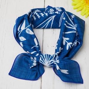 May include: A blue scarf with a white floral pattern is knotted in the center. The scarf is made of a lightweight fabric and has a dark blue border. Yellow flowers are in the background.
