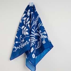 May include: A square, indigo blue cloth with a white botanical pattern of leaves, berries, and ferns. The cloth is hanging against a white wall, showcasing the detailed design. The edges are a darker shade of blue.