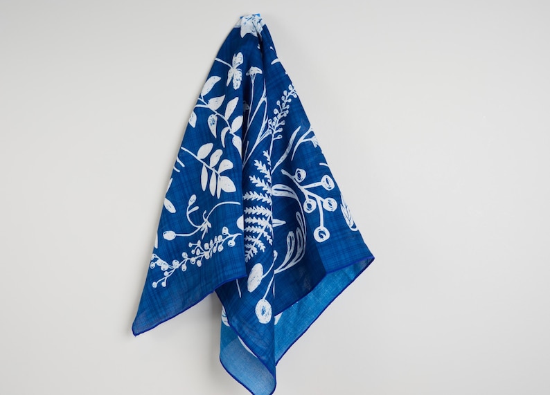 May include: A square, indigo blue cloth with a white botanical pattern of leaves, berries, and ferns. The cloth is hanging against a white wall, showcasing the detailed design. The edges are a darker shade of blue.