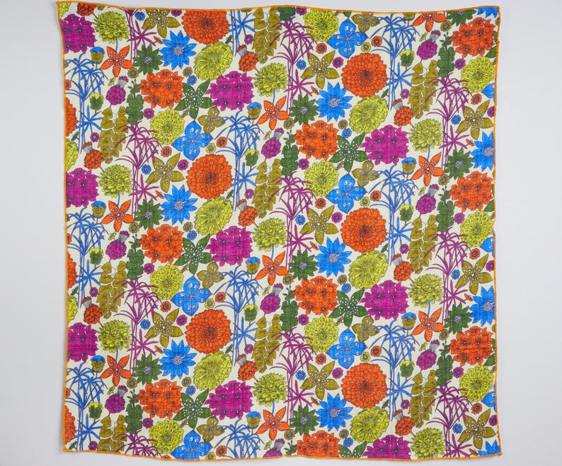 May include: A square textile with a vibrant floral pattern. The design features orange, pink, blue, and yellow flowers, along with green foliage, set against a cream background. The edges are trimmed with a thin orange border.