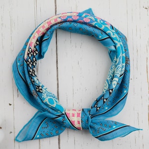 Könnte beinhalten: Ein leuchtend blaues Bandana mit komplizierten Mustern in Weiß, Rosa und Schwarz. Das quadratische Tuch ist geknotet und zeigt sein Design. Der Stoff ist leicht, perfekt als Hals- oder Kopfaccessoire.