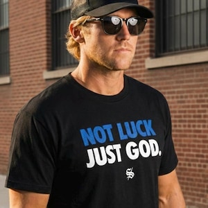 Puede incluir: Una camiseta negra con el texto "NOT LUCK JUST GOD" en azul y blanco. La imagen también muestra una gorra de camionero negra con las palabras "SOVEREIGN SHIELD" y un diseño de cruz, y gafas de sol.