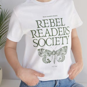 Könnte beinhalten: Weißes T-Shirt mit dem Text "READ BANNED BOOKS" über "REBEL READERS SOCIETY" in Grün. Darunter eine detaillierte Mottenillustration mit "EST. 1984". Das Shirt hat kurze Ärmel und einen Rundhalsausschnitt.