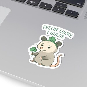 May include: A sticker featuring a cartoon opossum with a green shamrock and a matching headband. The text "FEELIN' LUCKY, I GUESS" is above the opossum. The sticker is on a laptop.