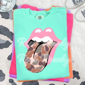 May include: A stack of colorful t-shirts. The top shirt is a light green with a pink lip graphic and a tongue with a brown and tan pattern. The text "Ree Rowdy" is written on the shirt. Other shirts are pink, orange, and purple.