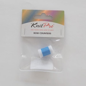 May include: A KnitPro row counter in a clear plastic retail package. The device is cylindrical with a bright blue center band and white ends. The packaging features a holographic top header with the text KnitPro FOR THOSE WHO LOVE TO KNIT.