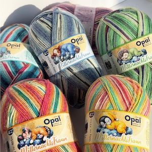 Puede incluir: Una colección de ovillos de lana de la marca Opal, disponibles en varias combinaciones de colores, incluyendo arcoíris, verde, azul y rosa. Cada ovillo tiene una etiqueta con el texto "Mitternachts Traum" y una ilustración de dibujos animados. Cada ovillo pesa 100g.