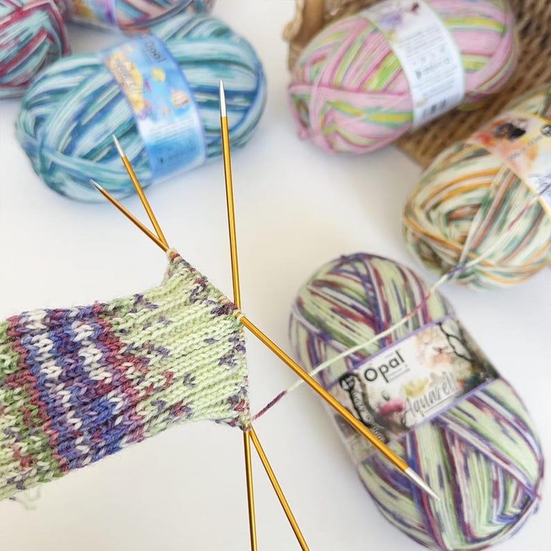 May include: Close-up of knitting needles with a partially knitted sock in progress. The sock features a green, purple, and white color scheme. Several balls of colorful yarn in shades of blue, pink, and green are scattered around the needles.