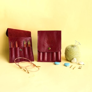 Könnte beinhalten: Zwei dunkelrote Lederetuis für Stricknadeln, eines offen und eines mit Druckknopf geschlossen, mit der Prägung lotus yarns. Enthält Rundstricknadeln, ein Knäuel hellgrünes Garn und kleine herzförmige Maschenmarkierer.