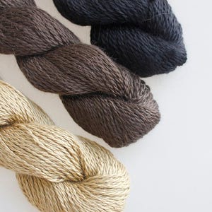 May include: Close-up of three skeins of yarn in varying shades of brown and black. The yarn is tightly twisted, showcasing its texture and color variations. The skeins are arranged diagonally, with a light beige, medium brown, and dark brown yarn.