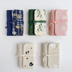 Puede incluir: Cinco estuches enrollables de tela en varios colores y patrones. Los estuches se cierran con un cordón a juego. Los diseños incluyen bordados florales, de hojas y de gatos. Los estuches miden aproximadamente 10 cm de ancho y 18 cm de alto cerrados.