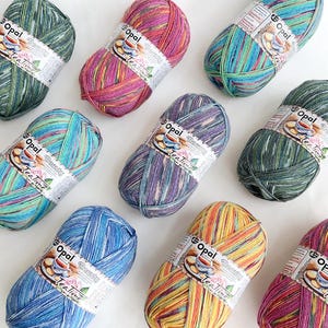 Op de afbeelding: Een assortiment kleurrijke Opal Beauty Tea Time garenbollen. Het garen is er in verschillende kleuren, waaronder groen, blauw, paars, roze en geel. Elke bol heeft een label met de merknaam en de tekst "Tea Time".