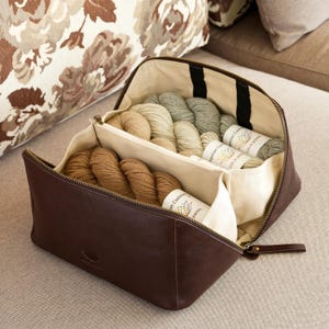 Può includere: Una borsa portaoggetti in pelle marrone scuro con un'ampia apertura a cerniera e un interno in tessuto color crema. La borsa è divisa in due scomparti, ciascuno riempito con diversi gomitoli di filato con etichette Lotus Yarns.