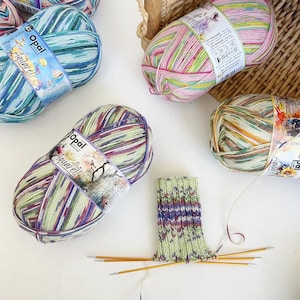 May include: Several skeins of Opal Aquarell yarn in various colors, including blue, green, purple, and pink. A partially knitted sock is shown with knitting needles. The yarn is 100g / 425m.
