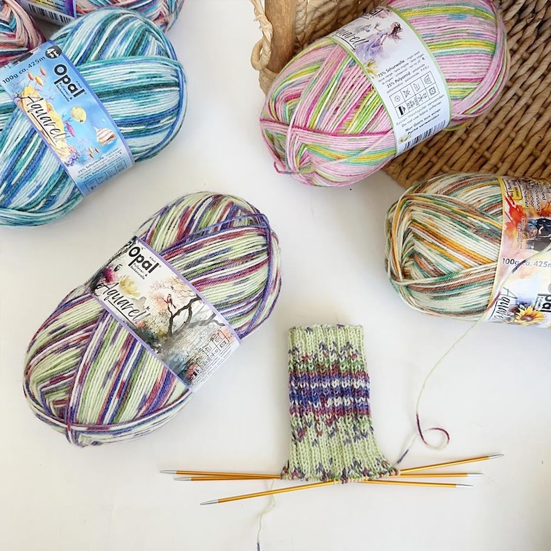 May include: Several skeins of Opal Aquarell yarn in various colors, including blue, green, purple, and pink. A partially knitted sock is shown with knitting needles. The yarn is 100g / 425m.