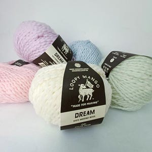 May include: A collection of Loopy Mango Dream yarn in various pastel colors, including pink, lavender, blue, and off-white. Each skein has a black label with the brand logo and text that reads "DREAM 100% MERINO WOOL".