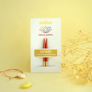 Puede incluir: Un paquete de agujas de tejer intercambiables Sahara de 9 cm. Las agujas son rojas con puntas doradas, y el embalaje presenta el logotipo de Lotus Yarns e información del producto. Un pequeño objeto decorativo está en primer plano.