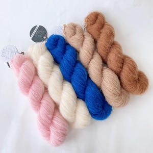 以下が含まれることがあります： ピンク、白、青、ベージュ、茶色の色合いのカラフルな糸の束のコレクション。 柔らかくふわふわした糸は、編み物や編み物のプロジェクトに最適です。 糸の束は白い表面に配置されています。