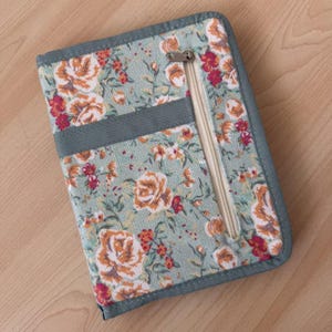 Pu&ograve; includere: Un portafoglio rettangolare in tessuto con sfondo verde chiaro e un motivo floreale di rose arancioni e rosse. Il portafoglio presenta una tasca verticale con cerniera color crema e una fascia in tessuto grigio tinta unita sul davanti.